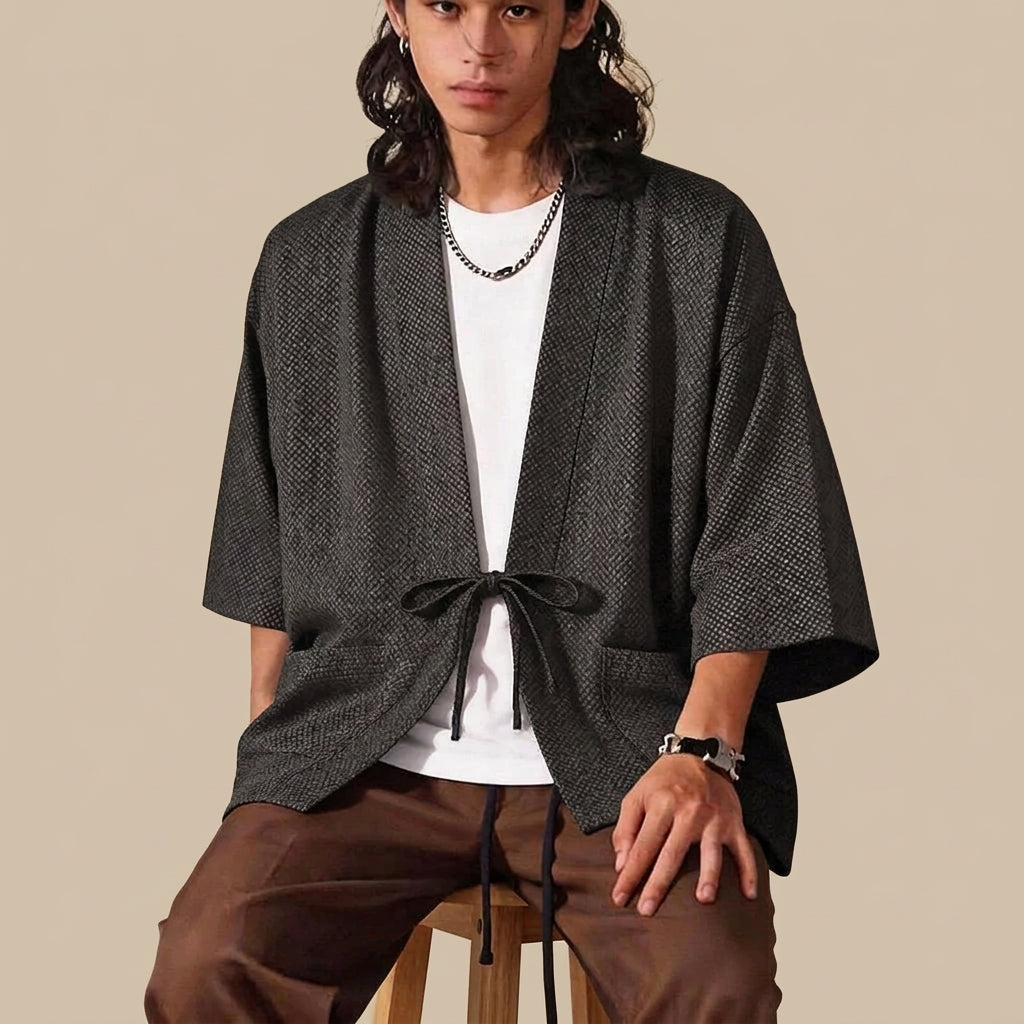 Man sitting on a stool wearing the Karo haori jacket in charcoal open over a white t-shirt, tied at the waist, with wide three-quarter sleeves